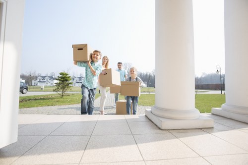 A group of four moving home, standing outside a residential property on a sunny day in Holland Park, London, with a view of houses and a street in the background. They are holding cardboard boxes of various sizes, with one woman in the foreground smiling and holding a box above her head as if ready to load it into a removals van or onto a trolley. The other three individuals are positioned behind her, also holding or carrying boxes, preparing to load items for a house move. The scene takes place on a paved area with large white columns and steps, suggesting a formal or historical building entrance, with the backdrop of a green lawn and trees. The image captures the process of house removals handled professionally by Holland Park Man with Van, emphasizing the careful handling of household belongings, such as boxes and small furniture, in a suburban London setting.