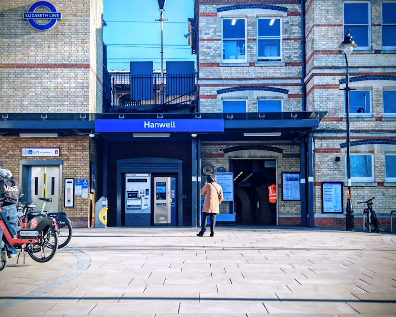 A woman dressed in a beige coat and black trousers stands outside the entrance of Hanwell station, a London Underground station with a brick facade featuring light and dark brickwork, blue signage, an