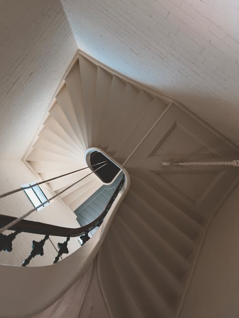 An interior view of a narrow spiral staircase inside a property, with white-painted wooden steps and black wrought iron balustrades featuring decorative elements. The staircase curves upward towards t